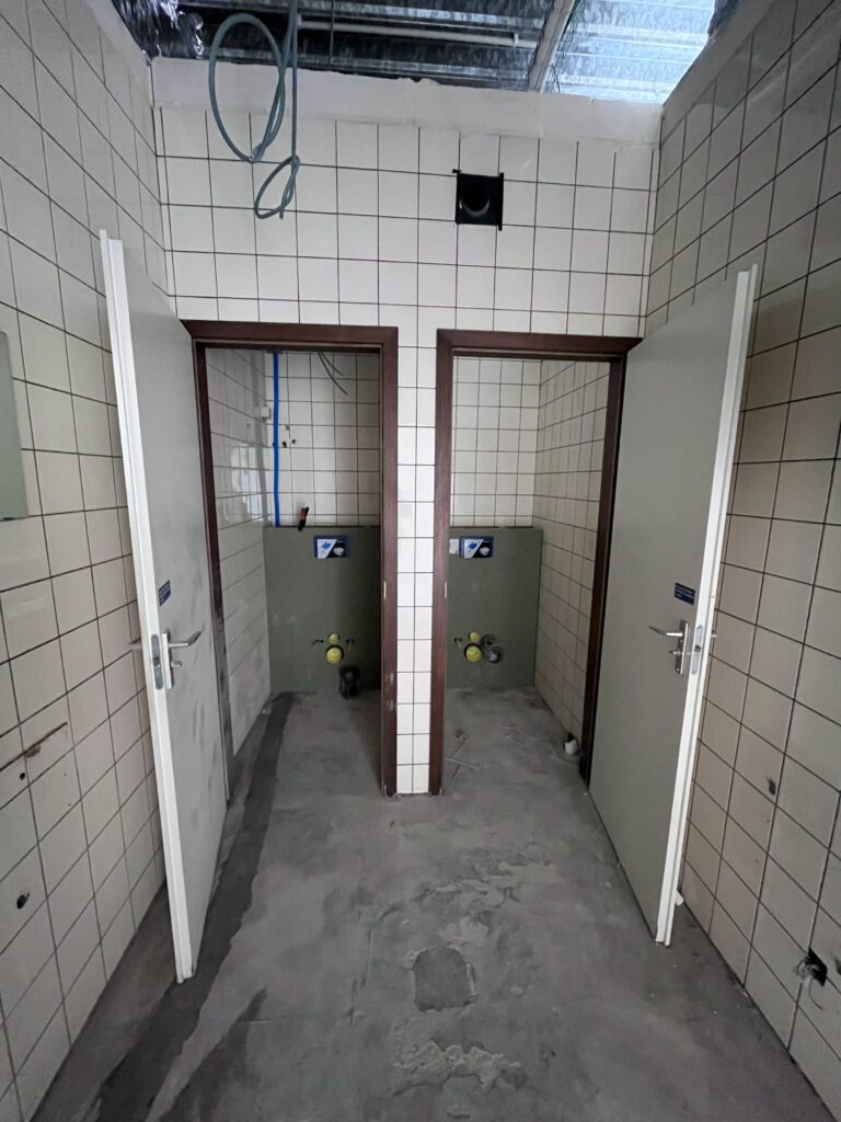 Verouderde sanitaire ruimte met betegelde wanden en vloer, gevoelig voor vuilophoping en slijtage.